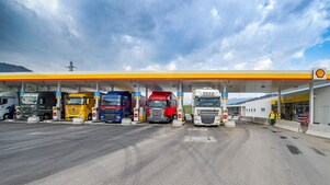 Gasolinera Shell con seis camiones aparcados. De izquierda a derecha: un camión blanco, verde oscuro, amarillo, azul oscuro, rojo y otro camión blanco.